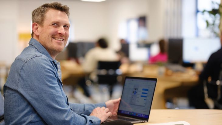 Andy is wearing a blue shirt and smiling at the camera, his laptop is in the background