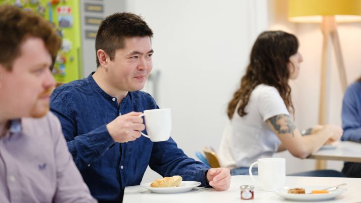 Dave drinking a coffee whilst talking to Alistair, there is a bacon sandwich on the table in front of both of them
