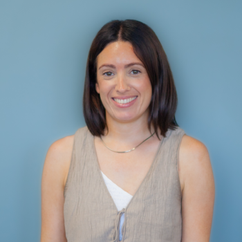 Emily has dark brown shoulder length hair. She is wearing a very light fawn coloured waistcoat style jacket over a white dress