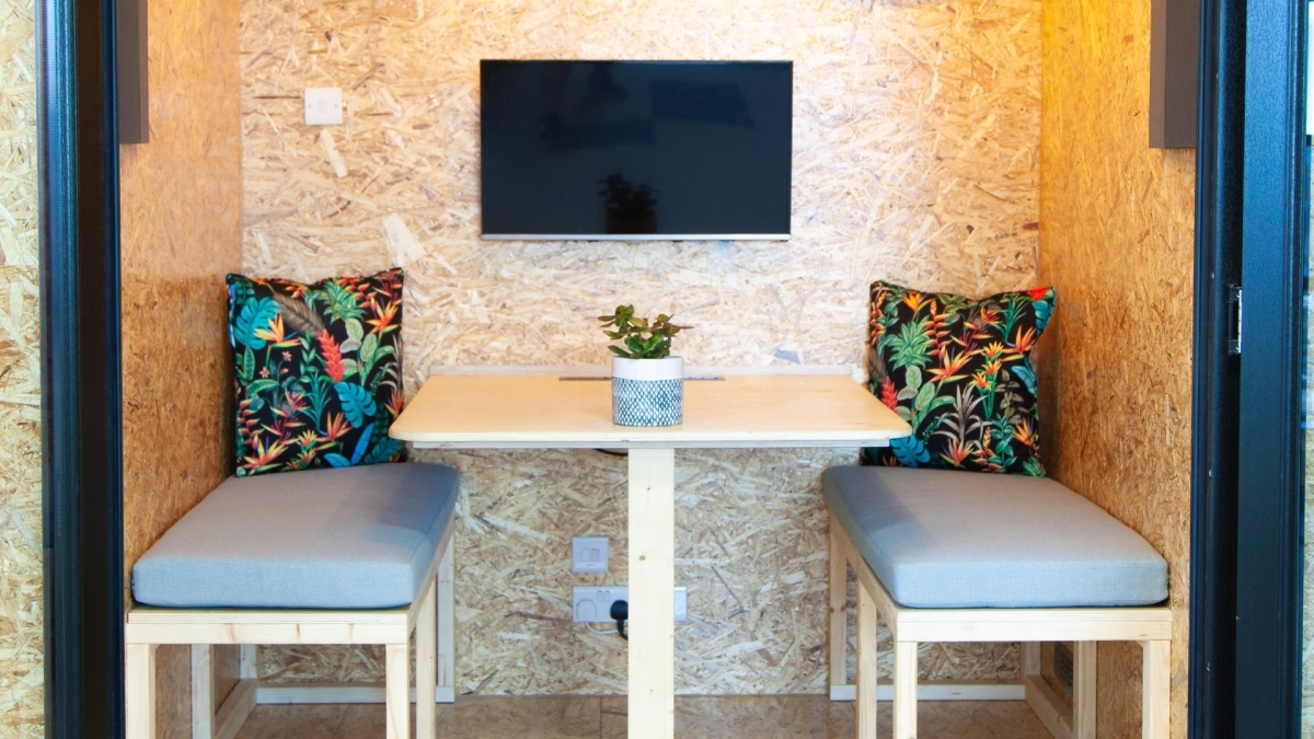 A photograph of one of the Brightec's meeting pods featuring; cushioned wooden benches, a wooden desk and a wall mounted monitor.